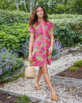 Model wearing a hot pink short-sleeved leopard-print dress with a button front, walking through a garden with a round woven handbag.