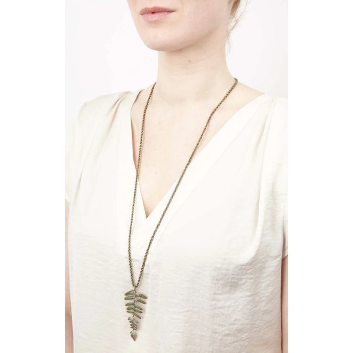 Long necklace with a leaf pendant worn by a person in a white top on a white background